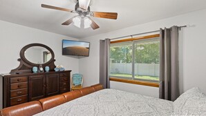Quarto com cama, penteadeira, espelho, ventilador de teto, janela com cortinas e um quadro na parede.