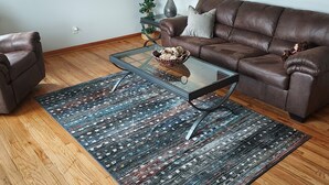 Uma sala de estar com um sofá de couro marrom, uma mesa de centro de vidro, um tapete estampado, uma televisão, um abajur e uma planta em vaso.