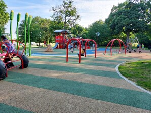 A playground with various play structures, including slides and swings, surrounded by trees and a clear sky. -  King Bed with fenced in backyard & Large 65" TV (Bourbonnais)
