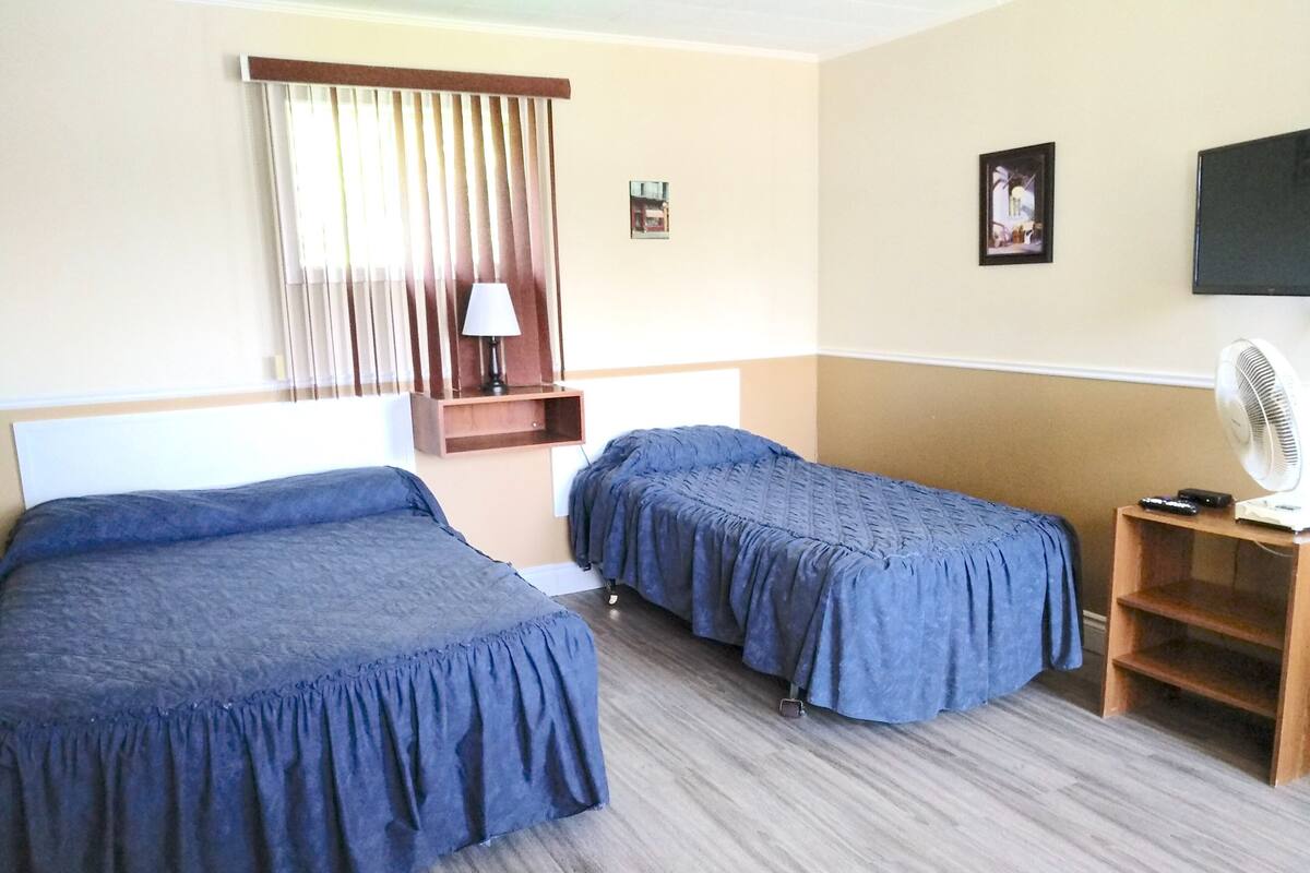 A hotel room with two single beds, a wooden nightstand with a fan, a small table, and a window with curtains.