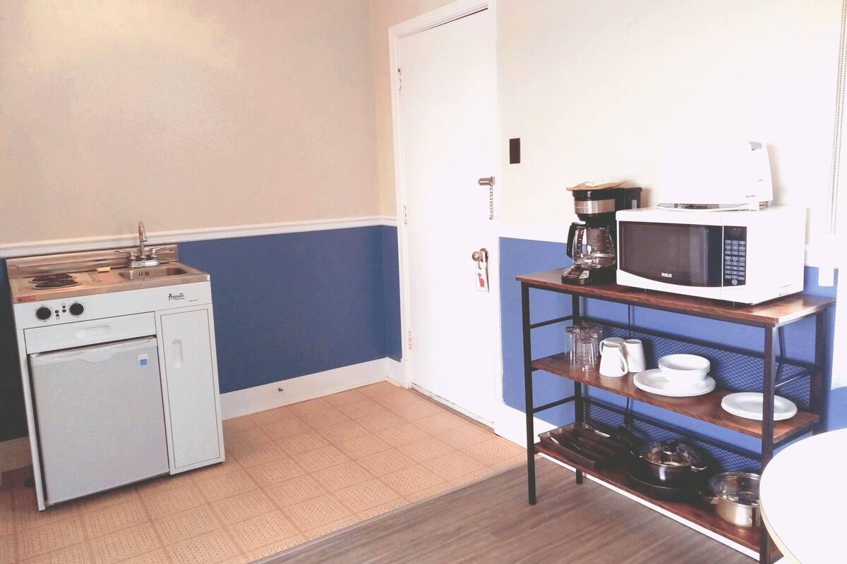 A compact kitchenette with a stove, oven, microwave, and a small table with dishes and a coffee maker.