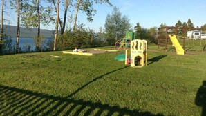 A grassy outdoor area with playground equipment, including a slide and climbing structures, near a body of water and trees. - Motel Fort Ramsay (Gaspe)