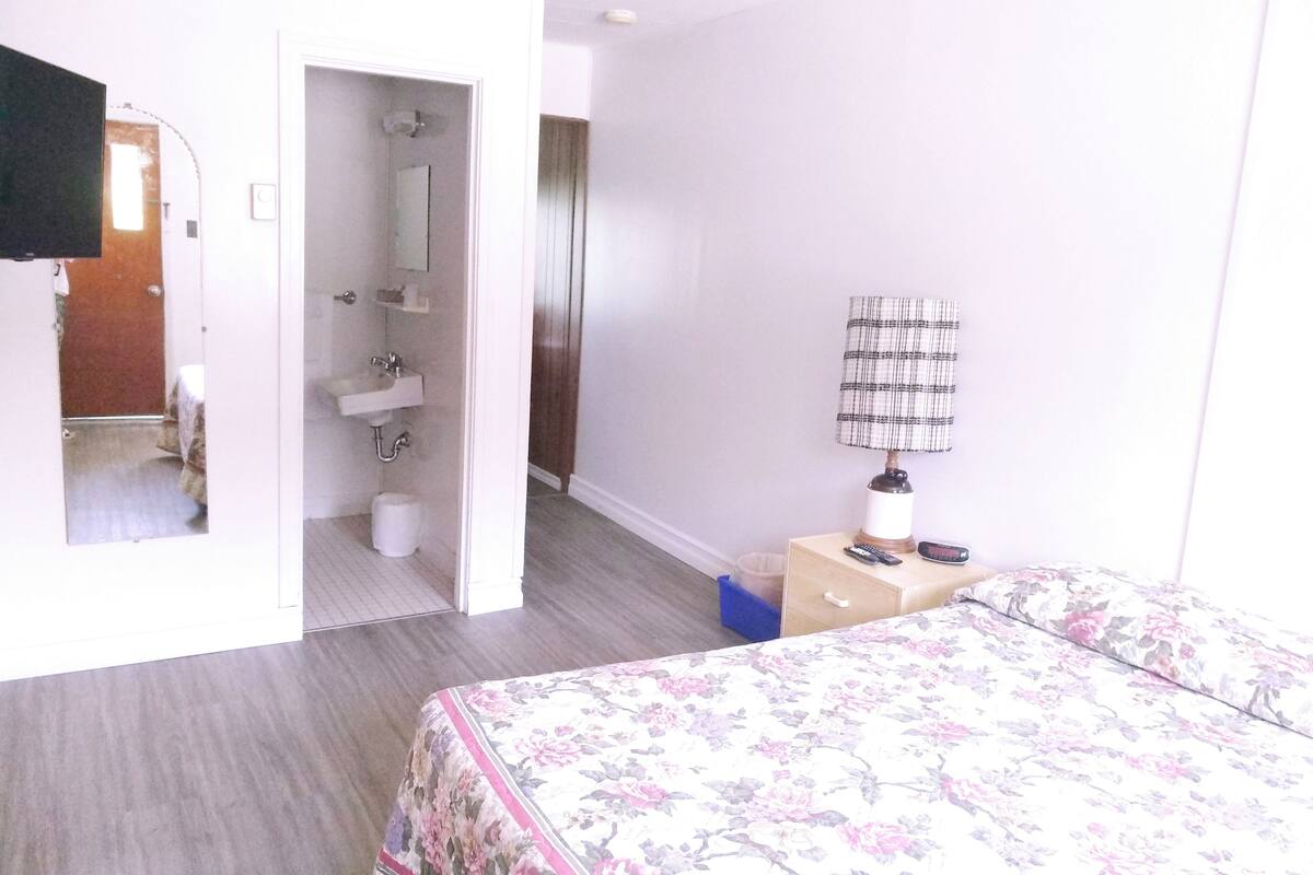 A hotel room with a bed, a bedside table with a lamp, a mirror, a bathroom with a sink and toilet, and a door leading to another room.