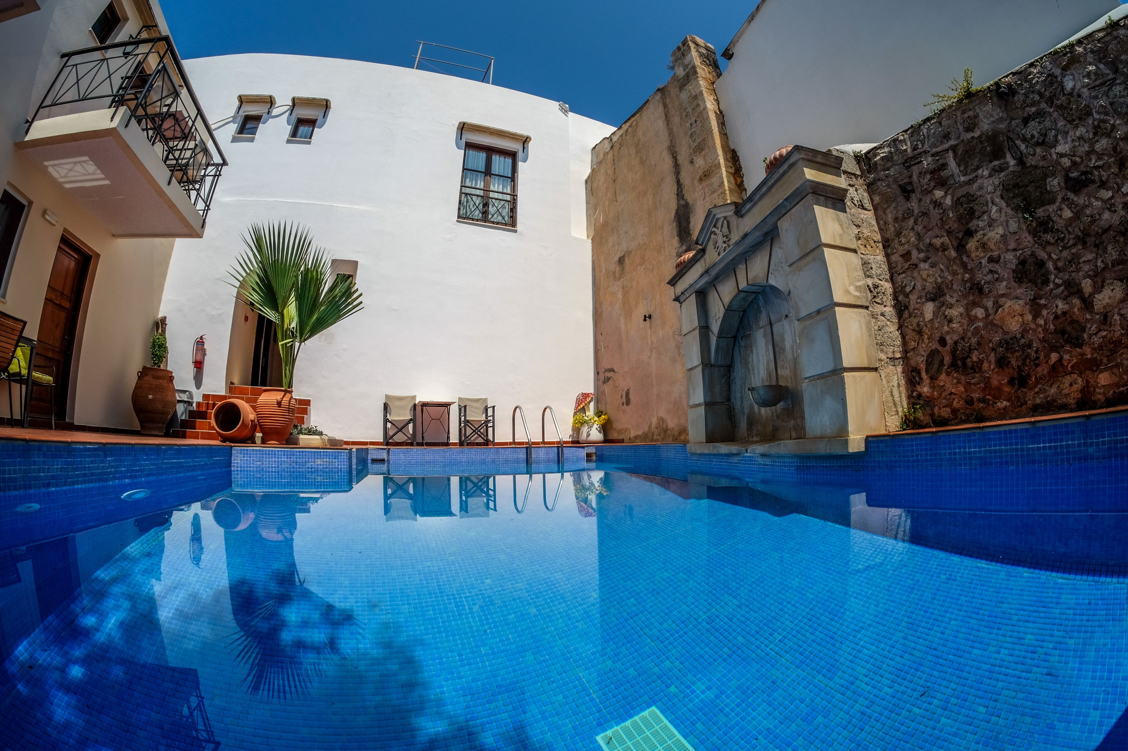 Ein Swimmingpool mit glatter, blauer Wasseroberfläche, umgeben von einem weißen Gebäude mit einer Steinmauer, einer Holztür und einem kleinen Balkon mit Metallgeländer.