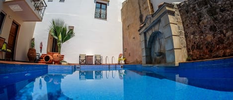 En swimmingpool med ett klart, blått vatten, omgiven av en vit byggnad med en stenmur, en trädörr och en liten balkong med ett metallräcke.