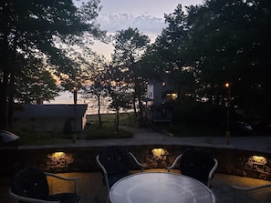 An outdoor patio area with a round table and chairs, a stone wall, and a view of a body of water and buildings in the background.