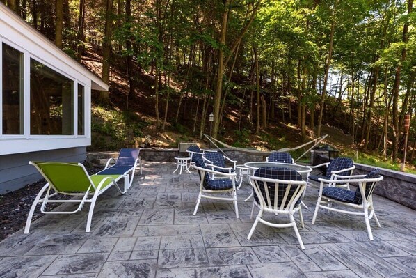 An outdoor patio with stone flooring, white wicker chairs with blue cushions, and a view of a forested area. - Beach View Cottage - Beautiful View of the Lake! (Holland)