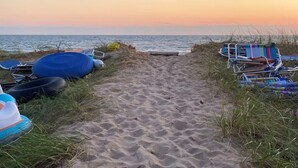 Un sendero arenoso que conduce al mar, con sillas de playa a la izquierda y una fila de sillas de playa a la derecha, además de un flotador azul.