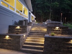 A set of stone steps leading to a house with a modern design, featuring a well-lit entrance and a small American flag on the right side. - Beach View Cottage - Beautiful View of the Lake! (Holland)