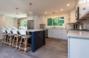 A modern kitchen with white cabinetry, a central island, and a dining area with wicker chairs. - Beach View Cottage - Beautiful View of the Lake! (Holland)
