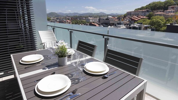Un balcone con un tavolo apparecchiato per due, vista su una città con edifici e montagne.