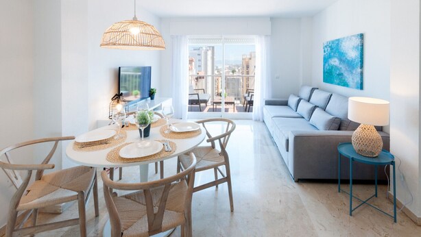 A modern living room with a grey sofa, a round dining table with wicker chairs, a hanging light fixture, and a view of the city through large windows.