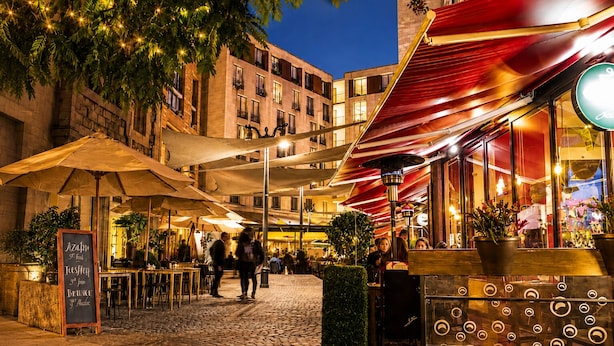 Un café al aire libre con mesas y sombrillas, rodeado de edificios.
