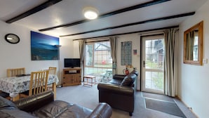 A cozy living room with a sofa, a TV, and a painting of a seascape. There is a dining table with chairs and a window with curtains. - 01 Tudor Court, Tolroy Manor (Hayle)