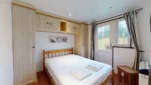 A bedroom with a wooden bed, white bedding, a small bedside table, a window with curtains, and a ceiling fan. - 01 Tudor Court, Tolroy Manor (Hayle)