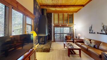 A cozy living room with a stone fireplace, wooden ceiling, and large windows.