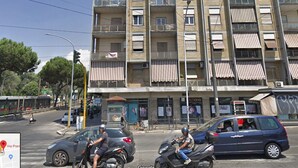 Un edificio de varios pisos con balcones, una calle con coches y un autobús, y un paso de peatones.