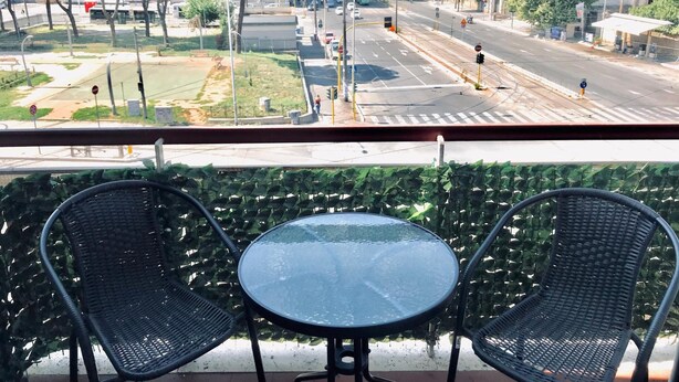 Un balcón con dos sillas de mimbre y una mesa de vidrio, con vistas a una calle urbana con edificios y árboles.
