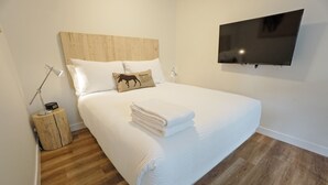 A neatly made bed with a white comforter and a folded white towel, a wooden headboard, a mounted flat-screen TV, and a small wooden nightstand with a lamp. - Basecamp Resorts Canmore I Micro-Suite, Dog Friendly (Canmore)