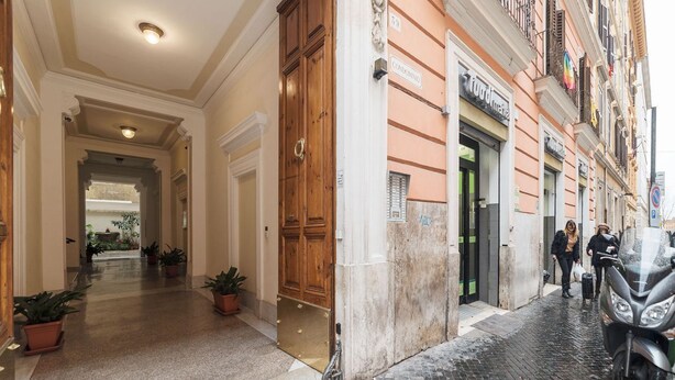 Entrada de hotel con puerta de madera, pasillo con plantas en macetas y calle con una motocicleta.