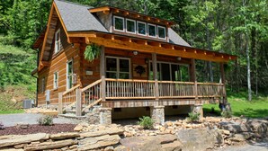A log cabin with a stone foundation, a wooden deck, and a large window on the front. - Riverbend Cabin! A Luxurious New Private Waterfront  Log Cabin. (Waynesville)