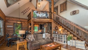 A rustic interior with a wooden staircase, a loft bedroom, a dining area, and a cozy living room with a fireplace. - Riverbend Cabin! A Luxurious New Private Waterfront  Log Cabin. (Waynesville)