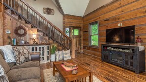 A cozy living room with a wooden staircase, a fireplace, a television, and a comfortable seating area. - Riverbend Cabin! A Luxurious New Private Waterfront  Log Cabin. (Waynesville)