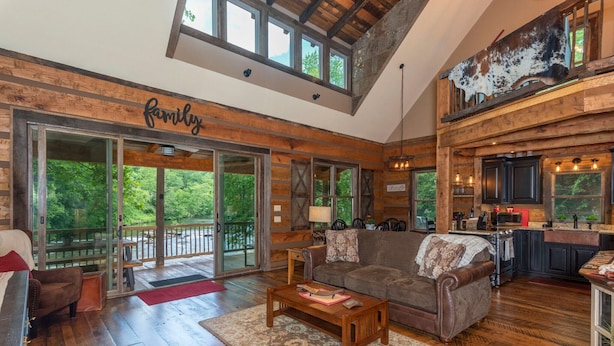 A spacious living room with a large couch, wooden flooring, and a view of the outdoors. - Riverbend Cabin! A Luxurious New Private Waterfront  Log Cabin. (Waynesville)
