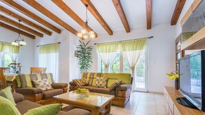 A spacious living room with wooden beams, a large flat-screen TV, and a dining area with a table set for four.