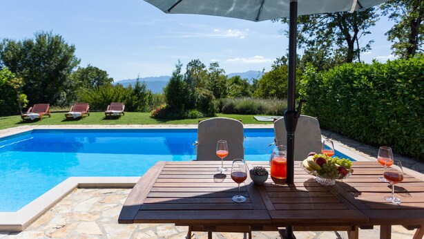 A poolside table with drinks and fruit, overlooking a swimming pool and garden chairs.