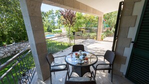A covered outdoor patio with a round table and four chairs, a stone wall, and a view of a swimming pool and greenery.