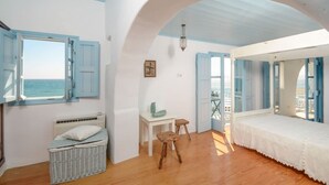 A room with a bed, a small table, a wicker basket, and a view of the sea through open shutters. - Casa Del Mare - Unique Beachfront Cycladic Villa @ Agios Prokopios (Agios Prokopios)