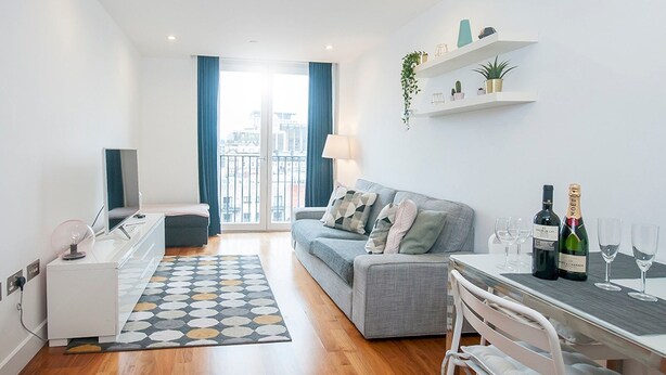 Um apartamento moderno com um sofá cinzento, uma mesa de jantar com copos de vinho, um pequeno armário branco e vista para a cidade através de grandes janelas.