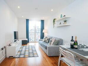 A modern apartment with a grey sofa, a dining table with wine glasses, a small white cabinet, and a view of the city through large windows.
