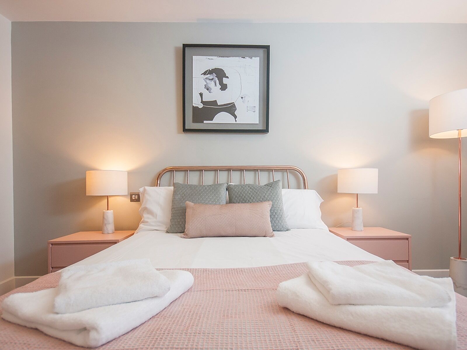 A neatly made bed with white and pink linens, two bedside tables with lamps, and a framed picture on the wall.