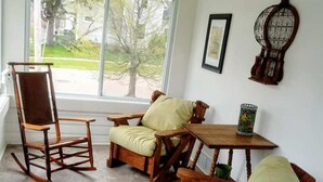 A room with a rocking chair, two cushioned chairs, a small table, and a window with a view of trees and a lawn. - Downtown, close to everything, family friendly, newly renovated. (South Haven)