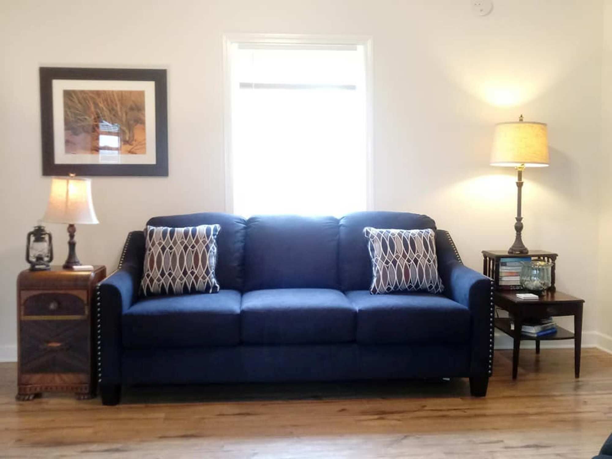 A room with a dark blue sofa, two patterned throw pillows, a wooden side table with a lamp, a framed picture on the wall, and a window with natural light.