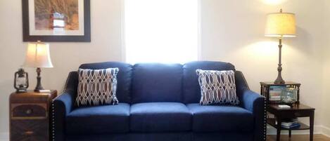 A room with a dark blue sofa, two patterned throw pillows, a wooden side table with a lamp, a framed picture on the wall, and a window with natural light.