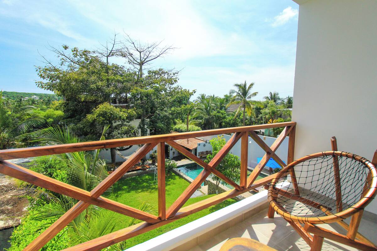 Un balcón con baranda de madera, una silla de mimbre y vistas a una piscina rodeada de vegetación exuberante y palmeras.