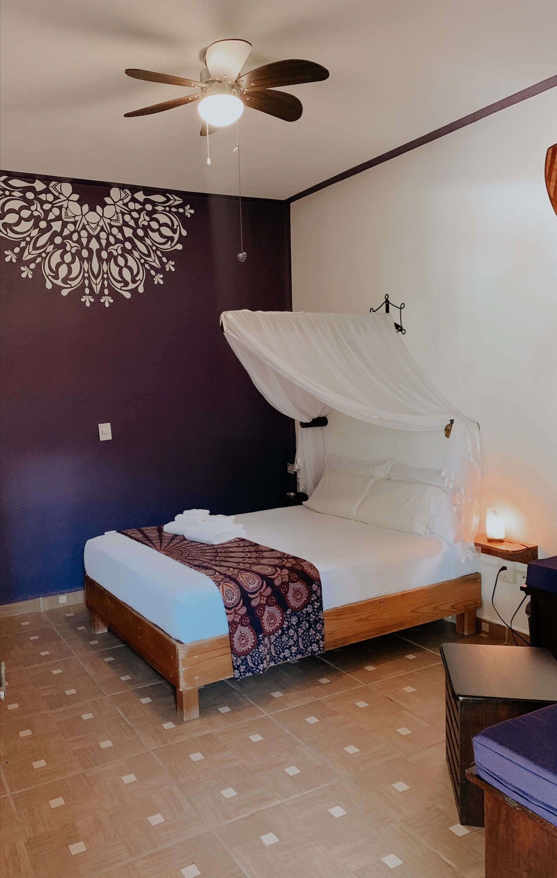 Un dormitorio con una cama bajo dosel, un ventilador de techo, una pared con diseño decorativo y una mesita de noche con lámpara.