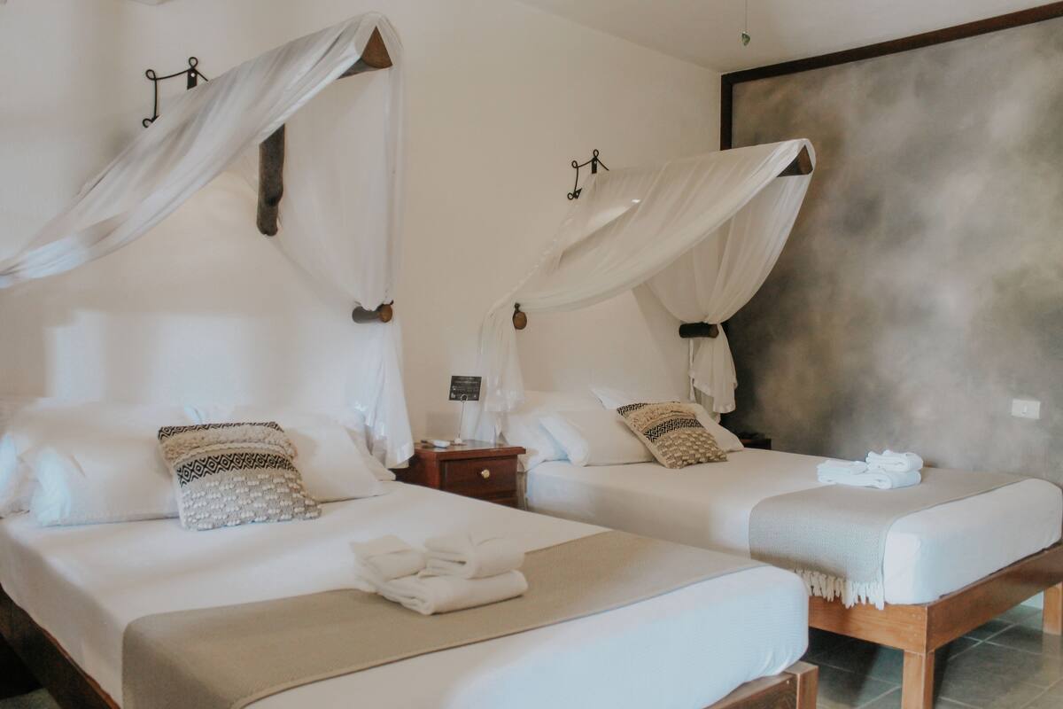 Habitación de hotel con dos camas, cada una con dosel, una mesita de noche con lámpara y vista a una pared con textura.