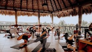 A yoga class in a room with a thatched roof, wooden floor, and a view of palm trees.