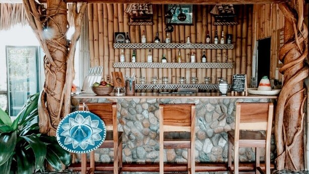 A rustic bar area with wooden shelves displaying bottles, a stone counter, and two wooden chairs.
