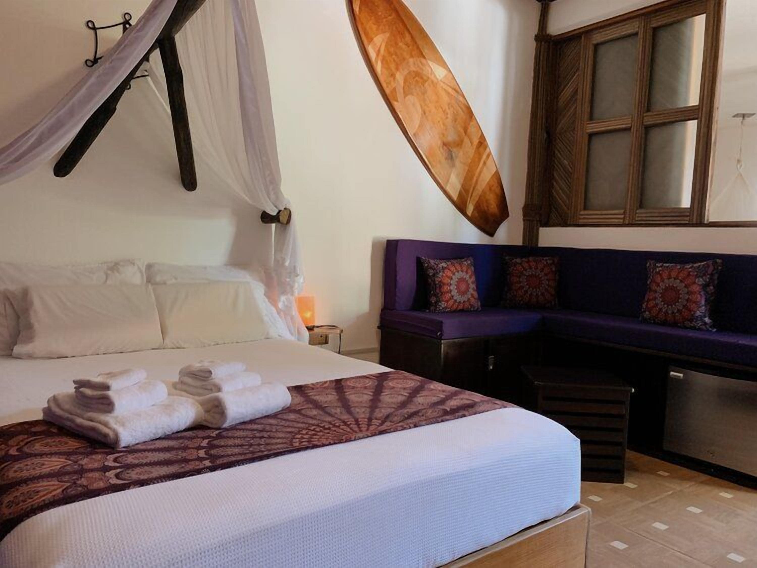 Uma cama com dossel, um banquinho com almofadas, uma cabeceira de madeira e uma janela com persianas.