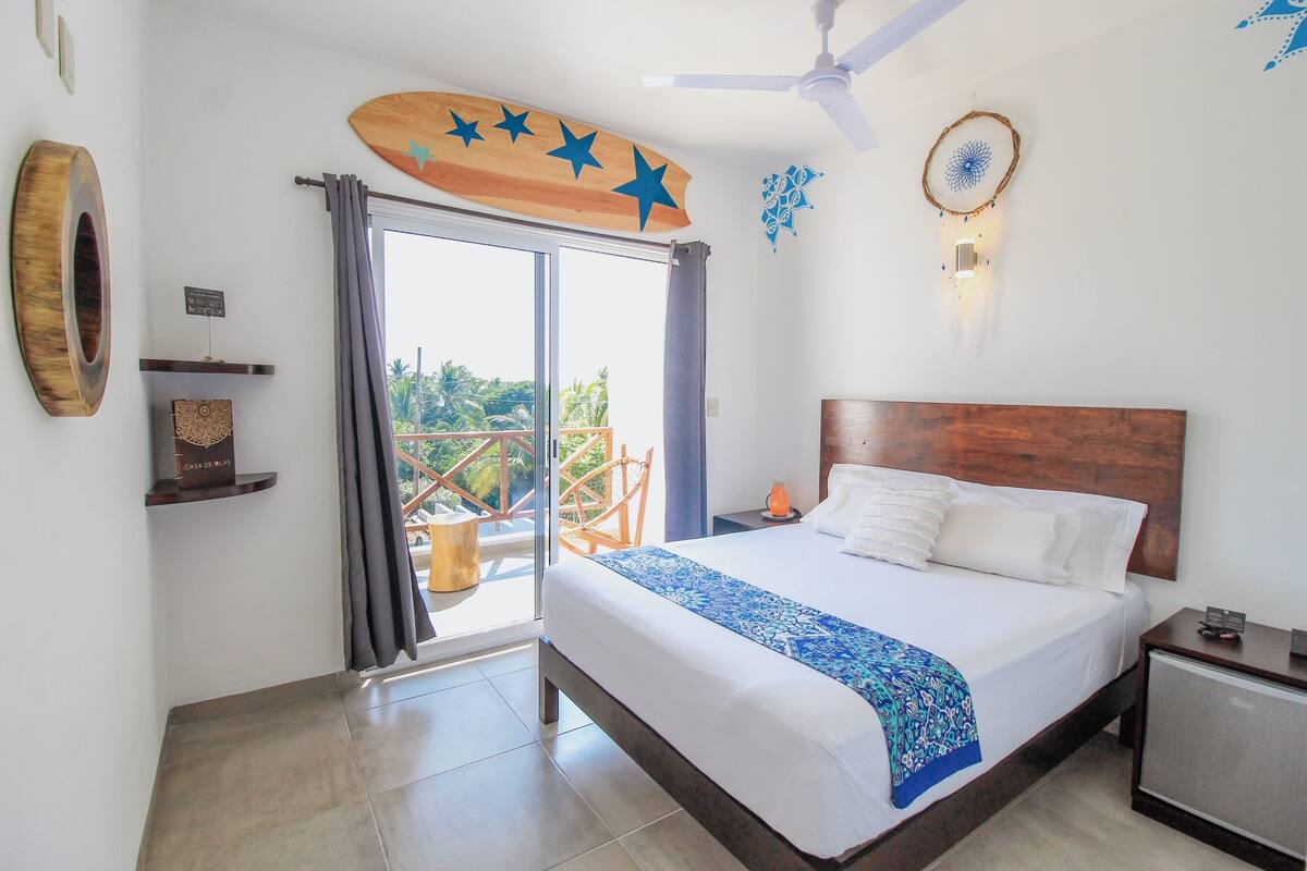 Habitación de hotel con cama, ventilador de techo, cabecero de madera, mesita de noche y ventana con cortinas. Incluye un balcón con una silla y detalles decorativos en la pared.