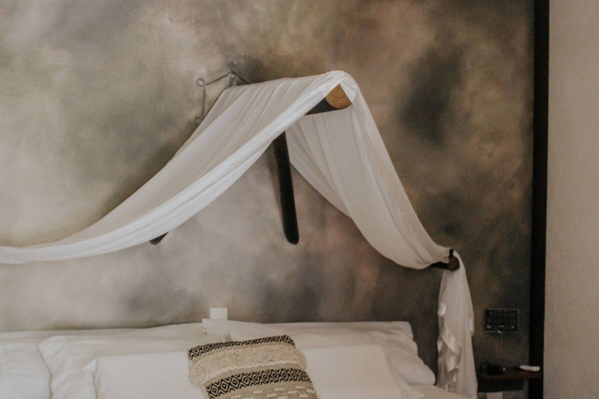Un dormitorio con cama, ropa de cama blanca, dosel, ventilador de techo y pared con textura.