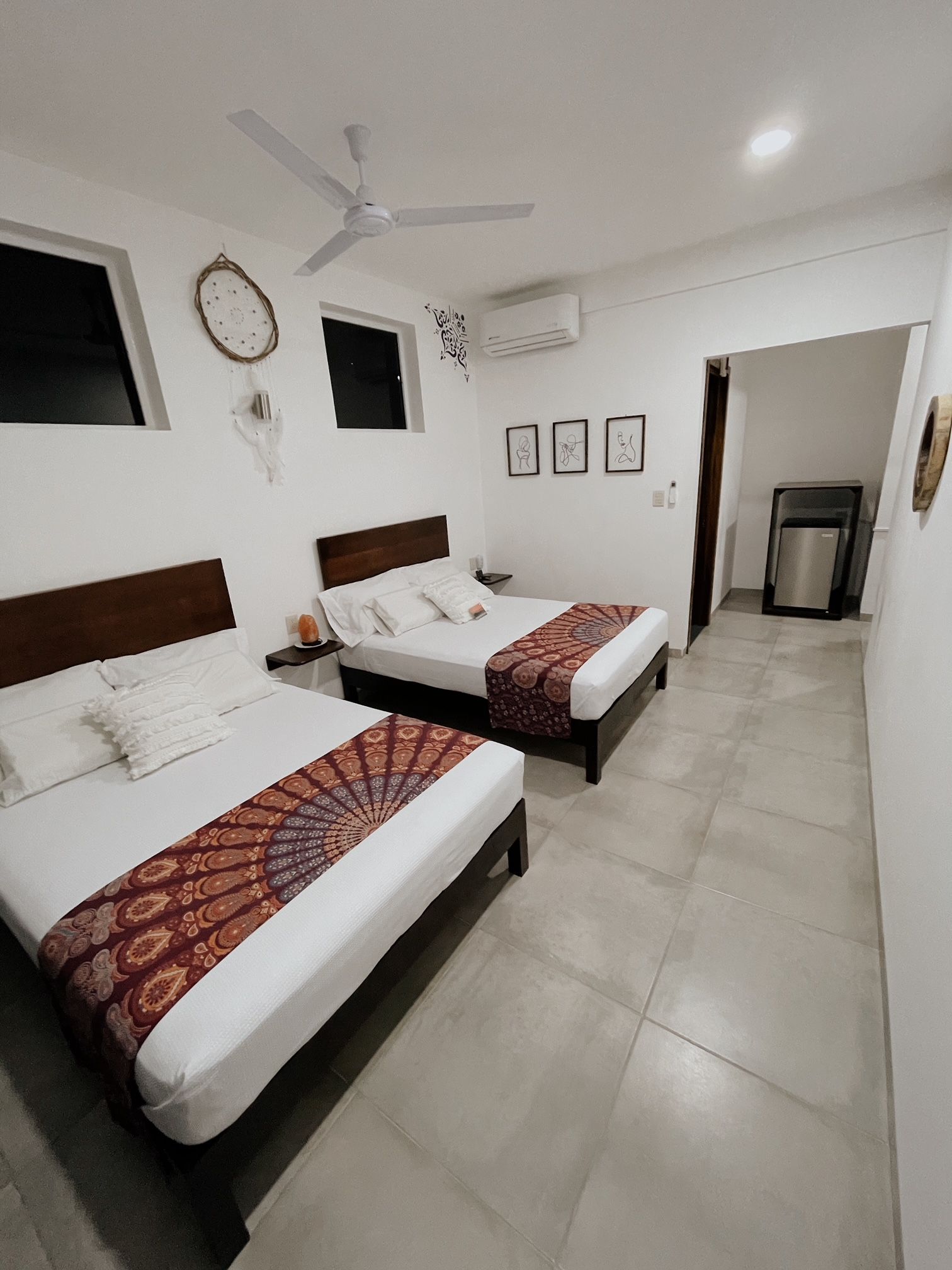 A hotel room with two beds, a ceiling fan, a small window, and a door leading to a kitchenette.
