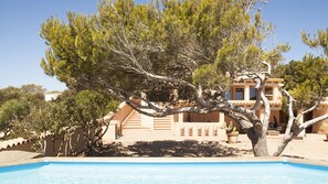 A poolside view of a Mediterranean-style villa with a large tree in the foreground, a swimming pool, and a building with a tiled roof and beige walls.