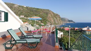 Una terraza con sillones de lona verde a rayas, una mesa con un paraguas de colores y vistas al mar y edificios en la colina.