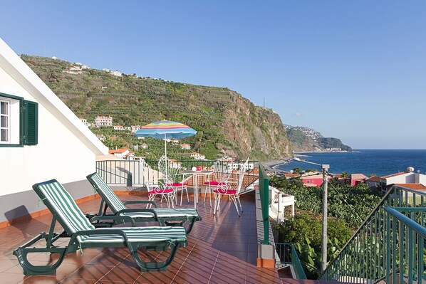 A terrace with green striped lounge chairs, a table with a colorful umbrella, and a view of the sea and hillside buildings.